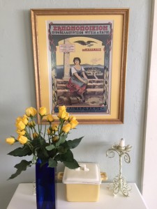 yellow roses in a black vase on a table with a Greek poster in the background.